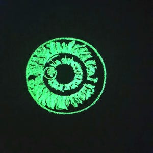 May include: A circular embroidered patch featuring a stylized human eye design. The iris and pupil are rendered in bright, glowing neon green thread against a solid black background, creating a high-contrast, detailed graphic representation.