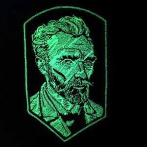 Può includere: Una toppa ricamata verde che brilla al buio con un ritratto di Vincent van Gogh. La toppa ha la forma di uno scudo.