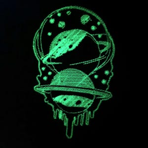 Może przedstawiać: Zielona, świecąca w ciemności haftowana naszywka przedstawiająca planetę z pierścieniami i rogal księżyca z gwiazdami. Planeta i księżyc kapie zielonym płynem.