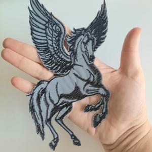 Peut inclure: Un écusson brodé gris à repasser représentant un cheval ailé, ou pégase, avec des points noirs.