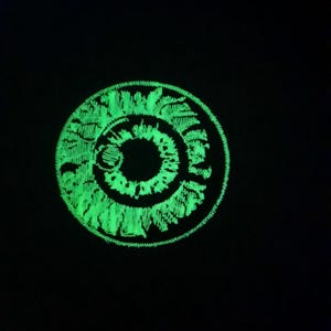 Puede incluir: Un parche circular verde que brilla en la oscuridad con un diseño de círculos concéntricos.