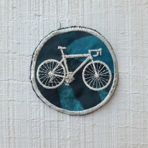 Pode incluir: Um remendo de bicicleta bordado branco em um fundo circular azul turquesa. O remendo tem uma borda branca.
