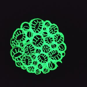 Può includere: Una toppa circolare caratterizzata da un intricato ricamo verde neon luminescente su uno sfondo nero uniforme. Il design presenta un motivo denso simile al pizzo con numerose piccole aperture ovali e circolari interconnesse.