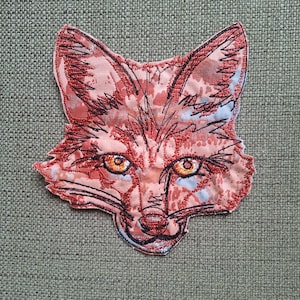 May include: A fabric patch shaped like a fox head featuring a printed pattern in shades of peach, orange, and white. The design is accented with black embroidered outlines defining the ears, eyes, whiskers, and snout, with bright yellow eyes.