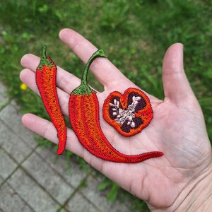 May include: Embroidered chili pepper patches in red and green. The image shows two whole peppers and one cross-section, all with detailed stitching. The patches are likely for clothing or craft projects.