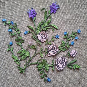 May include: An embroidered floral patch featuring three pale pink roses with dark outlines, surrounded by green stems and leaves, accented with small bright blue flowers and a single purple flower at the top, all stitched onto a textured fabric.