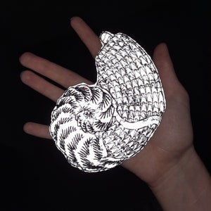 Puede incluir: Una pegatina reflectante de un animal enroscado con un diseño detallado y texturado. La pegatina es plateada y negra y tiene un acabado brillante e iridiscente.