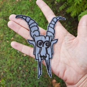 May include: A gray embroidered patch of a goat skull with black stitching. The goat skull has large horns and a detailed face.