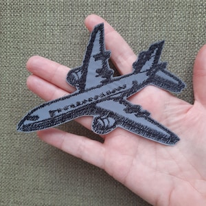 Peut inclure: Un écusson brodé représentant un avion gris avec des coutures noires. L'écusson est conçu pour ressembler à un avion vintage.