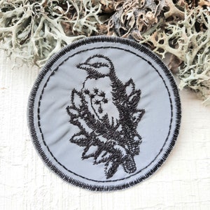Könnte beinhalten: Ein grauer gestickter Aufnäher mit einem schwarzen Vogel, der auf einem Ast sitzt. Der Vogel ist stilisiert und hat einen langen Schnabel und einen runden Körper. Der Ast ist mit Blättern verziert.