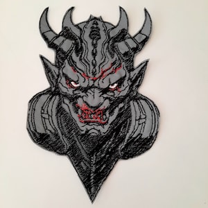 Puede incluir: Un parche bordado en negro y gris que representa la cara de un demonio con ojos y dientes rojos. El demonio tiene grandes cuernos y una expresión amenazante.