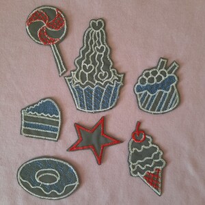 May include: A set of six iron-on patches featuring various sweet treats, including a cupcake, a lollipop, a donut, a star, a cake slice, and an ice cream cone. The patches are all gray with white and blue details, except for the star, which is red with white details.