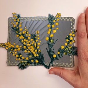 May include: Embroidered patch featuring a sprig of yellow mimosa flowers with green leaves on a gray background. The patch has a decorative stitched border and is approximately 4 inches wide.
