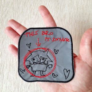 May include: A gray embroidered patch with a black line drawing of a person with a red circle around them. The text "THIS BRO IS INSANE" is above the drawing with an arrow pointing down.
