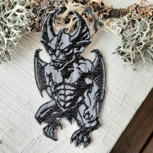 Könnte beinhalten: Ein schwarzer und grauer gestickter Bügeleisen-Aufnäher mit einer Gargoyle mit Flügeln, einem muskulösen Körper und einem bedrohlichen Ausdruck.