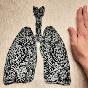 Puede incluir: Aplicación de encaje negro de pulmones y tráquea humanos. La aplicación presenta intrincados patrones florales y geométricos. El diseño se coloca sobre una superficie beige claro. Se incluye una mano para la escala.