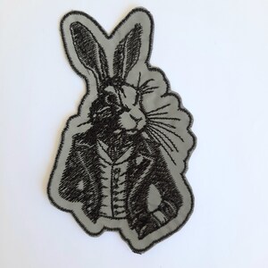 Op de afbeelding: Een grijs geborduurd patch met een zwart-witte illustratie van een konijn dat een jas en een strikje draagt.