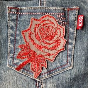 May include: A red embroidered rose patch with a grey outline, sewn onto a blue denim pocket. The patch is attached to a pair of jeans with a red stitched border. The label "KSQ" is visible on the right side of the image.