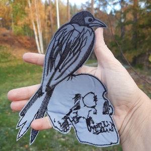 Op de afbeelding: Zwart-witte geborduurde patch van een kraai die op een schedel zit. De kraai heeft gedetailleerde veren en een scherpe snavel. De schedel heeft een gedetailleerde oogkas en tanden.