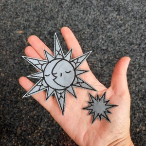 Puede incluir: Dos parches de fieltro gris con bordados negros. Un parche es un sol con una luna creciente en su interior. El otro parche es una estrella.