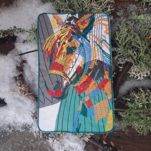 Pode incluir: Um remendo bordado colorido com uma cabeça de cavalo estilizada com um padrão geométrico. O cavalo é retratado em tons de vermelho, laranja, amarelo, azul e verde.