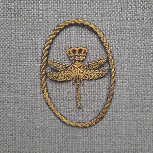 May include: A gold embroidered dragonfly with a crown on its head is centred in a black oval frame on a grey textured fabric.