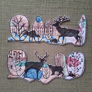 May include: Two embroidered patches with a forest theme. Each patch features a deer, trees, and a bear. The patches have a colourful design with blue, brown, and green hues. The patches are on a textured green background.