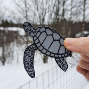 Peut inclure: Une applique de tortue de mer en feutre gris avec des broderies noires. La tortue est soulignée de fil noir et a un motif de carapace détaillé.