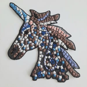 May include: A brown, blue, and white embroidered unicorn head patch. The unicorn has a long, spiraled horn and a detailed mane.