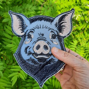 以下が含まれることがあります： ピンクの鼻と白いハイライトのある豚の顔の青い刺繍パッチ。豚は口を開けて笑っています。