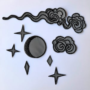 May include: A set of iron-on patches featuring a crescent moon, stars, and clouds. The patches are black and grey and have a reflective finish.