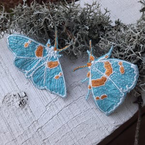 以下が含まれることがあります： オレンジのアクセントが付いた、青と白の刺繍が施された蝶が2匹。蝶は白い木製の表面に置かれています。