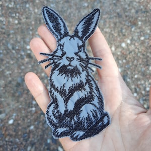 May include: A black and grey embroidered iron-on patch of a rabbit with long ears. The rabbit is sitting with its front paws tucked under its chin.