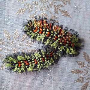May include: Two decorative, embroidered caterpillar-like shapes. The central body is black with orange and yellow accents. The edges are fringed with black and green threads. The background is a light gray fabric with gold and silver snowflake designs.