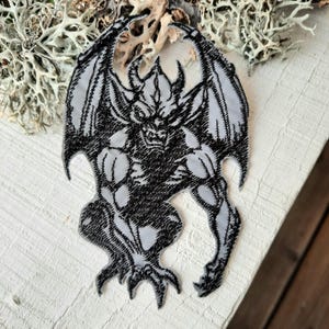 Könnte beinhalten: Ein gestickter Patch in Schwarzweiß, der eine Gargoyle mit Flügeln zeigt. Die Gargoyle hockt mit ausgebreiteten Flügeln.