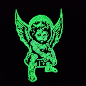 Può includere: Ricamo di un cherubino verde neon con ali, aureola e dettagli fini, su sfondo nero. Il cherubino è raffigurato in posizione seduta, con le braccia incrociate. Il ricamo ha un aspetto strutturato.