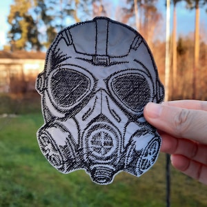 Op de afbeelding: Een geborduurde patch in zwart en wit van een gasmasker. De patch is gemaakt van stof en heeft een gedetailleerd ontwerp.