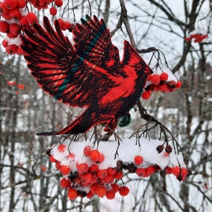 May include: A red cardinal bird with black markings is perched on a branch covered in snow. The bird is made of fabric and has a detailed design. Red berries are visible on the branch.