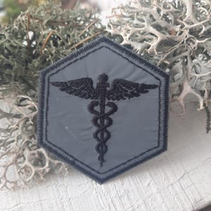 Puede incluir: Un parche hexagonal negro y gris con un símbolo médico bordado en negro, una serpiente enroscada alrededor de un bastón con alas.