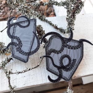 Può includere: Due toppe di tessuto grigio con disegni ricamati neri di linee e forme vorticose. Le toppe sono cucite sui bordi.