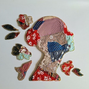 May include: A fabric collage in the shape of a human head profile, constructed from various patterned textile scraps including floral, lace, and solid fabrics in red, blue, pink, and brown, accented with gold-colored decorative stitching.