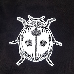 Peut inclure: Image brodée en noir et blanc d'une coccinelle sur un fond de tissu noir.