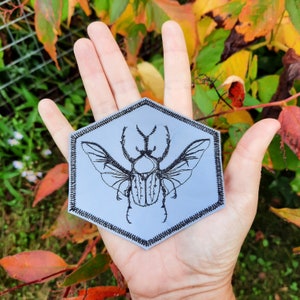May include: A gray hexagonal patch with a black embroidered image of a beetle with wings spread out.