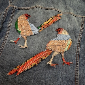 Pode incluir: Dois patches bordados de faisões coloridos sobre um fundo de denim azul. Os patches são detalhados com linhas intrincadas e sombreamento.