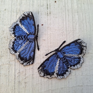 以下が含まれることがあります： 2つの刺繍された蝶のパッチ。蝶は、黒と白のディテールが施された詳細な青い翼を持っています。パッチは、衣類やアクセサリーに追加するのに最適な、テクスチャード加工された外観をしています。