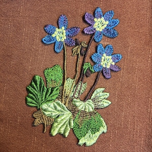 以下が含まれることがあります： 黄色い中心と緑の葉を持つ、青と紫の花が3つ刺繍された花の模様。 刺繍は茶色の布の背景に配置され、詳細なステッチと自然なテーマを示しています。