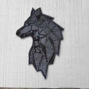 Pode incluir: Um remendo bordado com a cabeça de um lobo geométrico em preto e branco. A cabeça do lobo está virada para a esquerda e tem um design detalhado.
