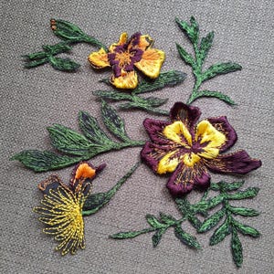 May include: An embroidered patch featuring three flowers with deep purple and bright yellow petals, accompanied by detailed green leaves and stems. The embroidery uses varied thread textures to create depth and color contrast across the floral design.