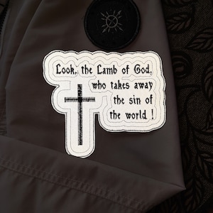 Può includere: Toppa ricamata con la scritta "Look, the Lamb of God, who takes away the sin of the world!" e una croce nera. La toppa ha un bordo bianco e si trova su uno sfondo grigio.