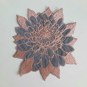 Pode incluir: Um remendo bordado de flor em cinza e ouro rosa. A flor é um design estilizado com um centro grande e muitas pétalas.